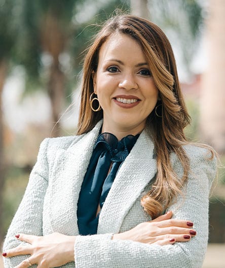Médica sorridente, vestindo blazer claro sobre blusa azul escura, com acessórios dourados, de braços cruzados, em ambiente ao ar livre.