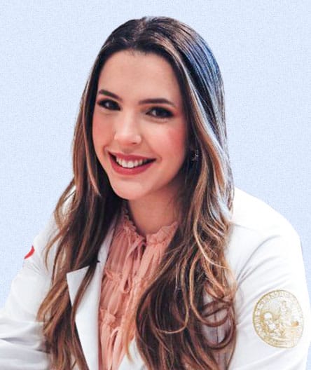 Médica sorridente com cabelos longos, vestindo jaleco branco com detalhes dourados no braço e blusa rosa delicada.