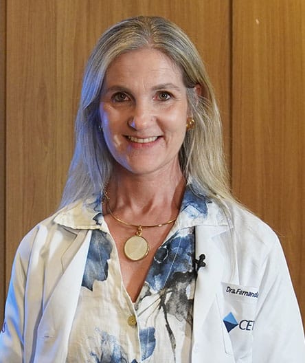 Médica sorridente, vestindo jaleco branco com logotipo do Cetrus, camisa estampada em tons de azul e colar dourado, em frente a um painel de madeira.