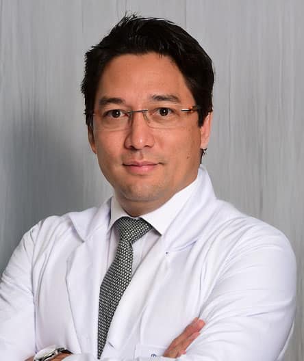 Dr. Marco Demange, vestindo jaleco branco e gravata cinza, com expressão confiante e braços cruzados, em frente a um fundo neutro.