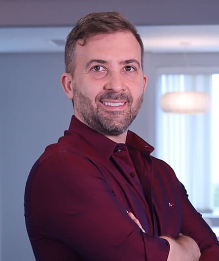 Médico sorridente, vestindo camisa social bordô, de braços cruzados, em ambiente moderno e iluminado.