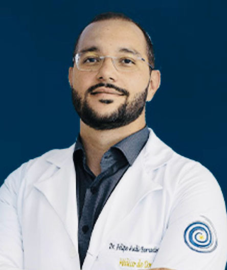Dr. Felipe Audi, médico especialista em dor, coordenador acadêmico, usando jaleco branco e óculos, posando com os braços cruzados diante de um fundo azul.