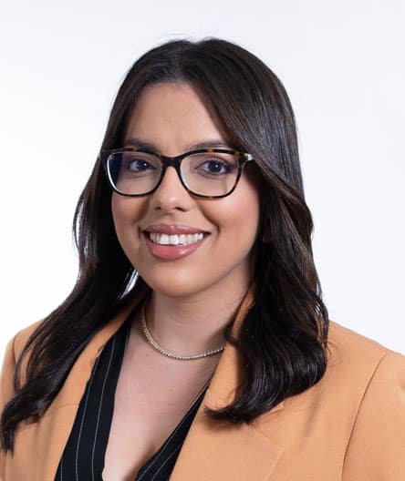 Retrato profissional de uma mulher jovem com cabelos castanhos escuros e lisos, usando óculos de armação escura, blazer bege e blusa preta, sorrindo em fundo branco.