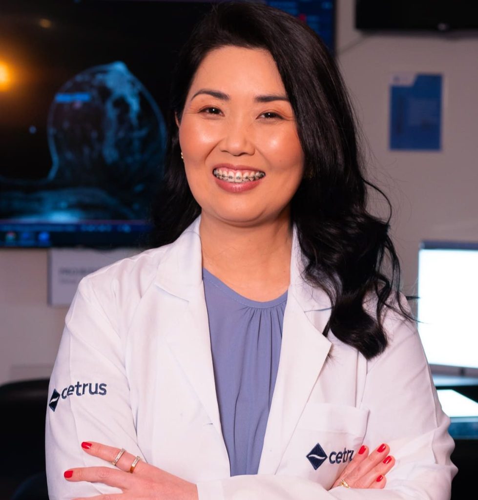 Médica radiologista usando jaleco do Cetrus, sorrindo e com os braços cruzados em uma sala de diagnóstico por imagem, com exame de ressonância magnética mamária ao fundo.