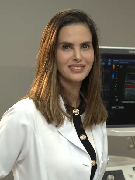 Profissional de saúde usa jaleco branco e sorri em frente a um aparelho de ultrassonografia em ambiente clínico.