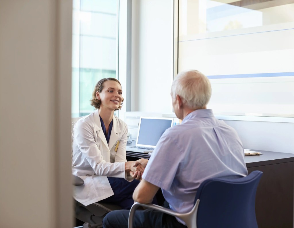 Profissional de saúde cumprimenta um paciente idoso em consultório médico moderno, demonstrando acolhimento e empatia.